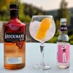 Et glass med T&T med appelsinskall på et bord med grønn og sommerlig bakgrunn. På hver side av glasset, til venstre flasken med Brockmans Orange Kiss Gin - til høyre Artisan Pink Sitrus Tonic. Til sammen blir dette: Sitrus-GT med Brockmans Gin Orange Kiss